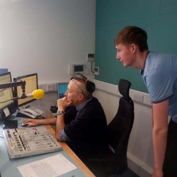 A radio station with a man sat at microphone and a young man looking behind his shoulder.